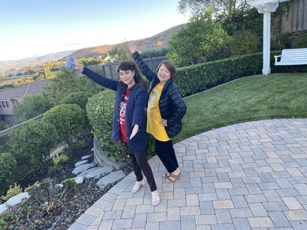 Two women posing happily outdoors on a paved patio with greenery and hills in the background.