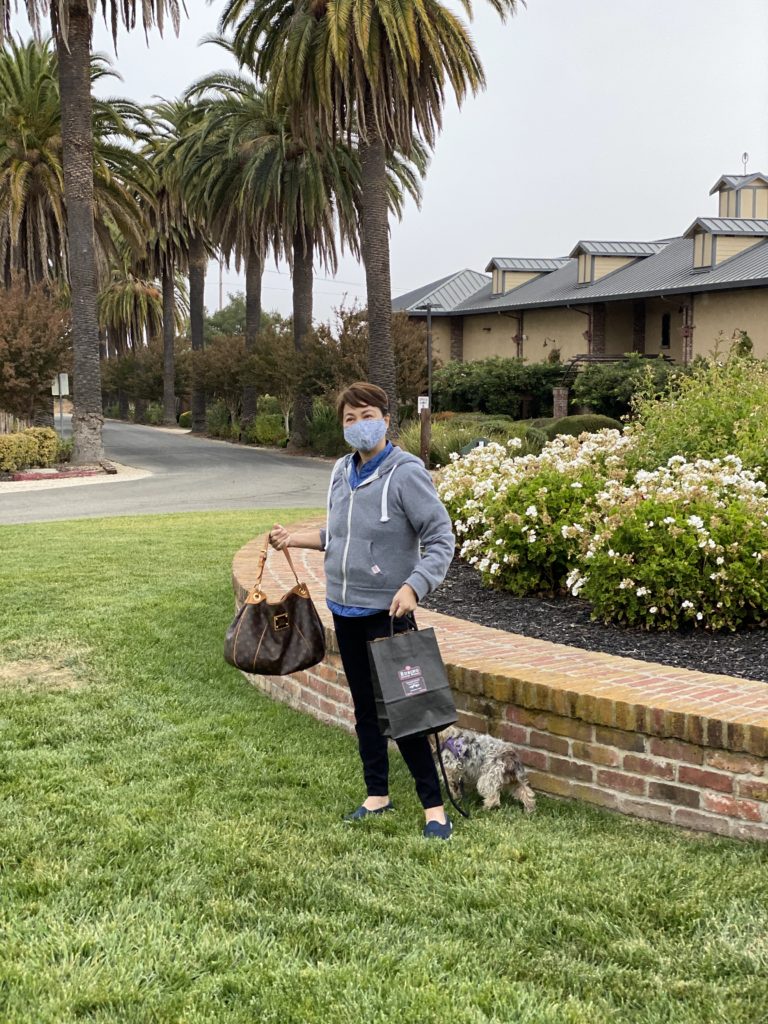 Person wearing mask carrying bags near brick planter and palm trees.