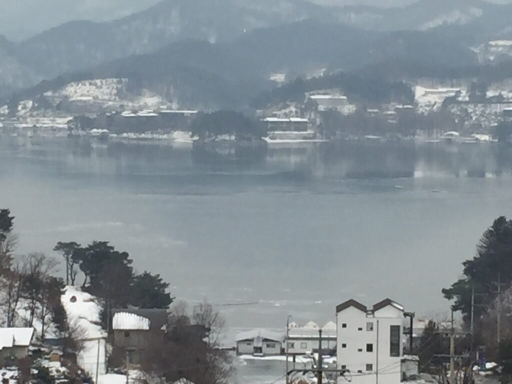 Snow-covered houses by a calm lake with distant mountains.