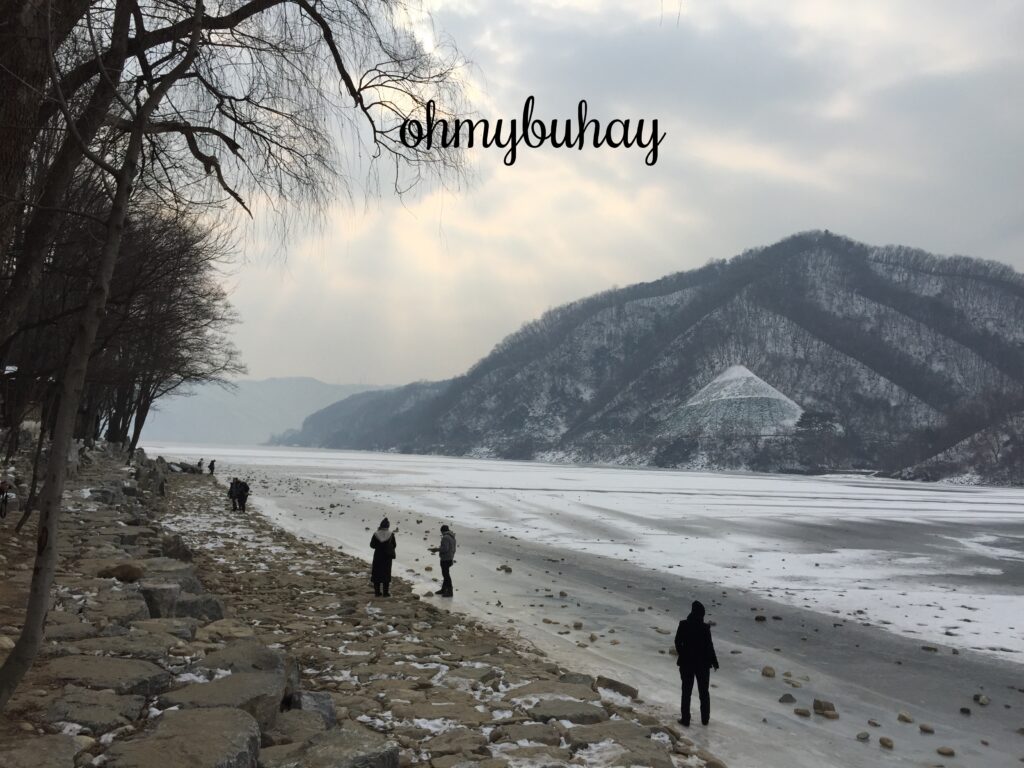 People walking along a rocky riverbank under cloudy skies.
