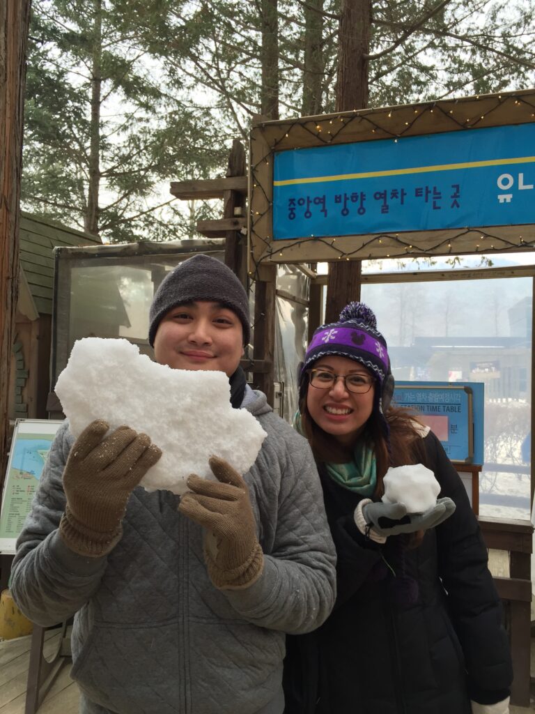 Two people smiling, holding large snowballs in a snowy outdoor setting.