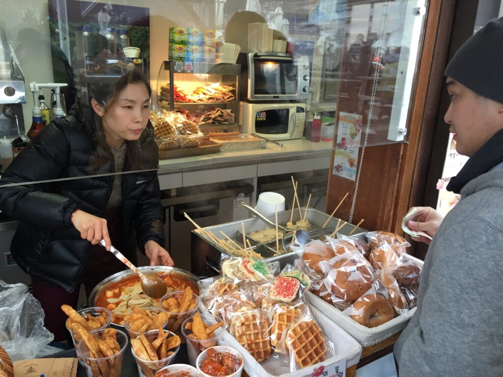 Woman serving food at a street stall with various dishes and snacks.