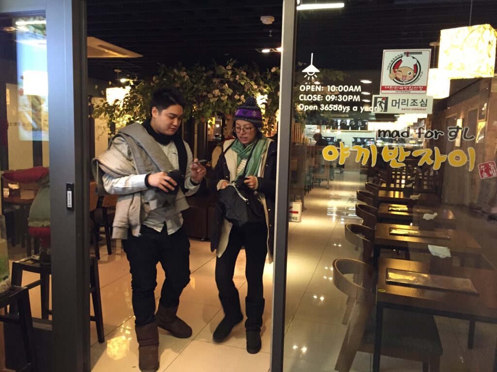 Two people standing and looking at their phones inside a cafe.
